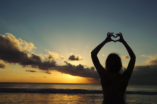 Elleri günbatımı arka plan karşı bir kalp şeklinde oluşturma genç bir kadın silüeti - Fotoğraf, Görsel