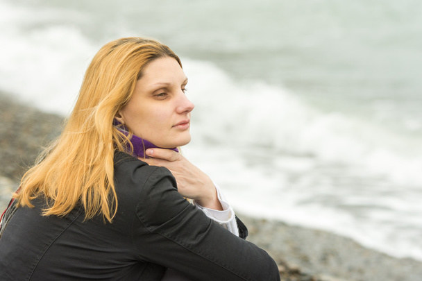 Una chica se sienta en la playa en un día nublado en clima frío y mirando pensativamente a la distancia
 - Foto, imagen
