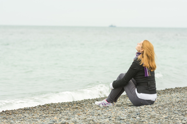Jeune fille assise sur une plage de galets face à la mer face à la brise de la mer par une journée nuageuse
 - Photo, image