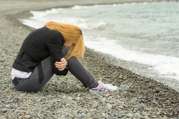 Chica en un día fresco en la playa se sienta pensativamente con su cara enterrada en sus rodillas
 - Foto, imagen