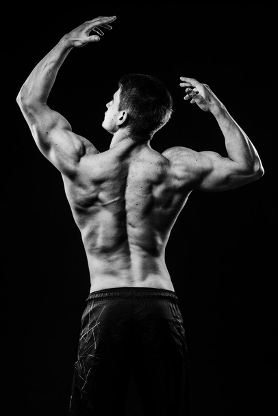 Young and fit bodybuilder athlete demonstrates biceps back view isolated on black background - Photo, Image
