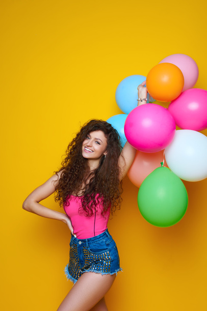 Young and beautiful curly girl in a pink shirt and blue shorts on a yellow background holding colorful balloons and laughing - Photo, Image