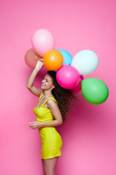 Jong en mooi krullend meisje in een gele jurk op een roze achtergrond houdt van kleurrijke ballonnen - Foto, afbeelding