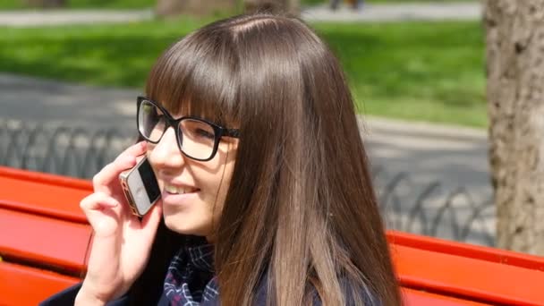 Молода жінка в окулярах говорить по скромному телефону в міському парку. Дівчина сидить на червоній лавці навесні і виступає на смартфоні. Крупним планом
 - Кадри, відео