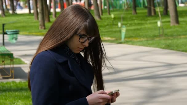 Kaunis silmälasipäinen nainen käyttää kännykkää ulkona puistossa - yksityiskohdat. Nuori viehättävä onnellinen tyttö rentoutuu kaupungin puistossa ja käyttää matkapuhelinta. Hän näyttää onnelliselta ja tyytyväiseltä.
 - Materiaali, video