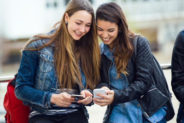 Two beautiful students using mobile phone in the street. - Zdjęcie, obraz