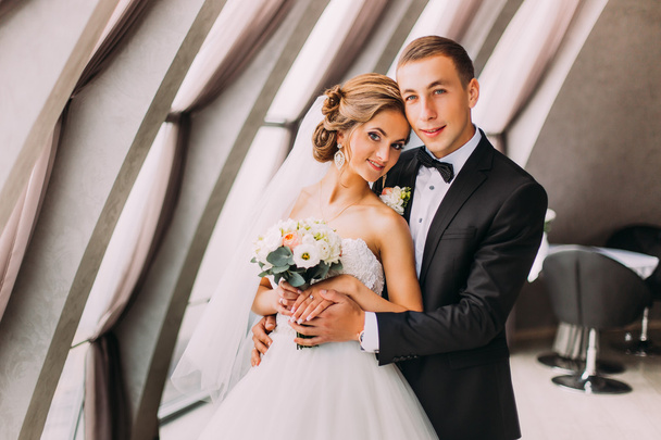 Gorgeous happy stylish groom hugs from behind beautiful bride on the luxurious interior background, close-up - Фото, изображение