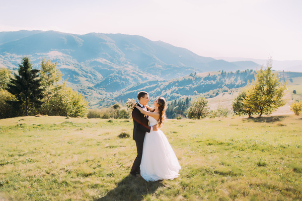 De bruid en bruidegom knuffelen op de weide met berg achtergrond. - Foto, afbeelding