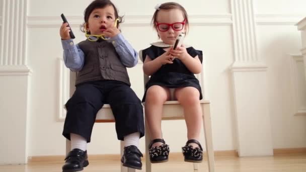 Baby boy and baby girl in glasses sit with mobile phones - Filmati, video