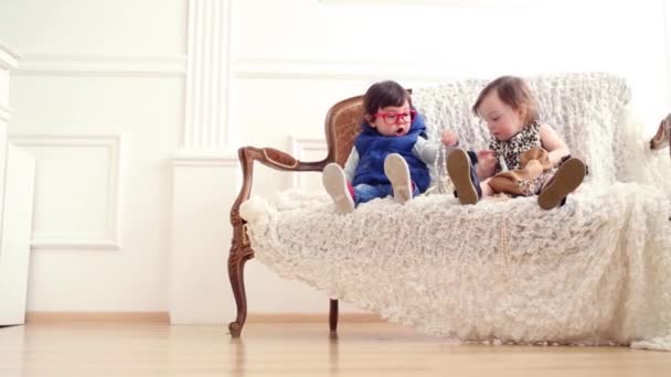 Beautiful baby girl in dress and baby boy sit on couch - Imágenes, Vídeo
