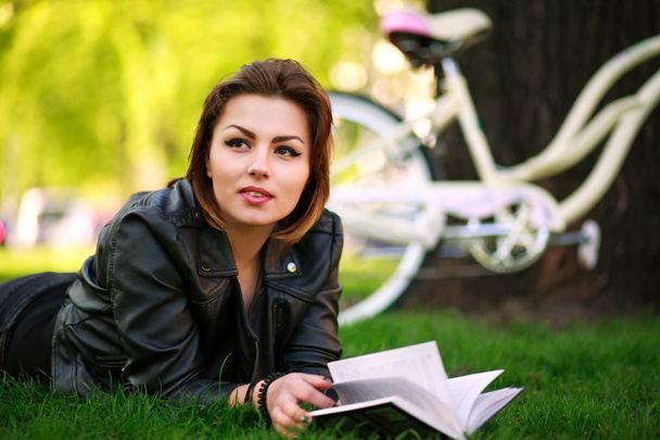 Young woman with bicycle reading book in city park on the grass - Photo, Image