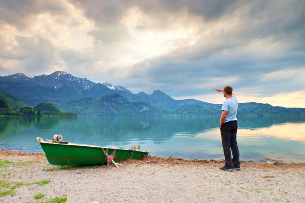 Hombre adulto en camisa azul caminar en el viejo barco de remo de pesca en la costa del lago montañas
. - Foto, Imagen