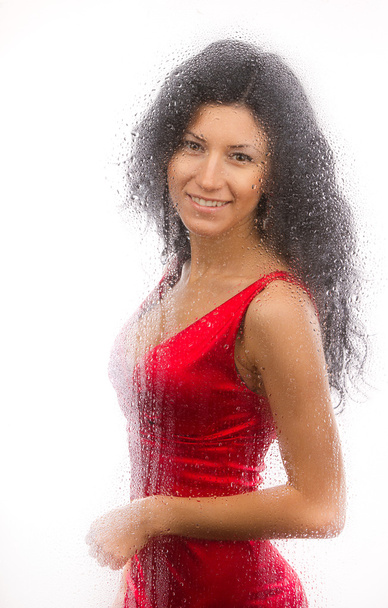 schönes Mädchen in rotem Kleid, hinter einem nassen Glas auf weißem Hintergrund stehend. - Foto, Bild