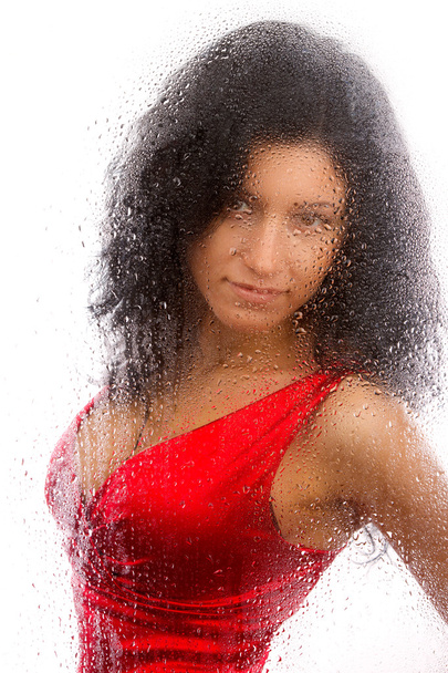 Beautiful girl in a red dress, standing behind a wet glass on a white background. - Φωτογραφία, εικόνα