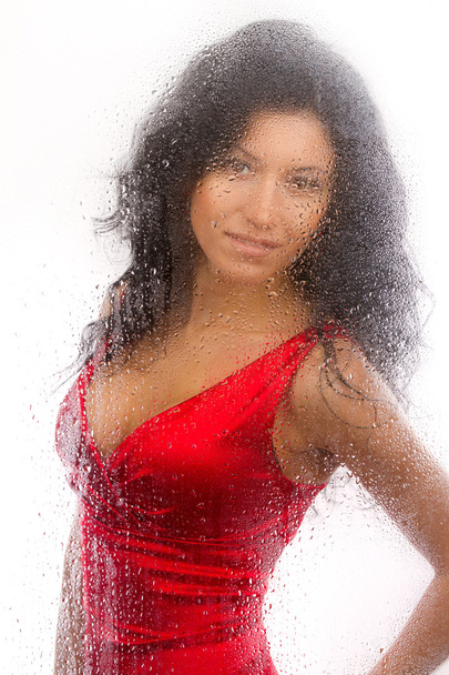 Beautiful girl in a red dress, standing behind a wet glass on a white background. - 写真・画像