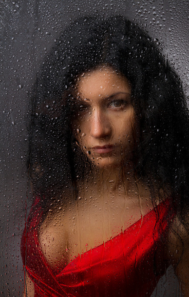 Beautiful girl in a red dress, standing behind a wet glass on a dark background. - Photo, Image