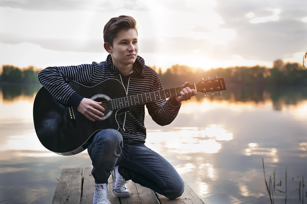 Young guy in a hood plays guitar on the bridge in the evening against the backdrop of a sunset on the river - Foto, imagen