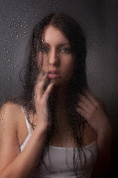 A beautiful girl in a white t-shirt, on a black background behind the glass. - Фото, зображення