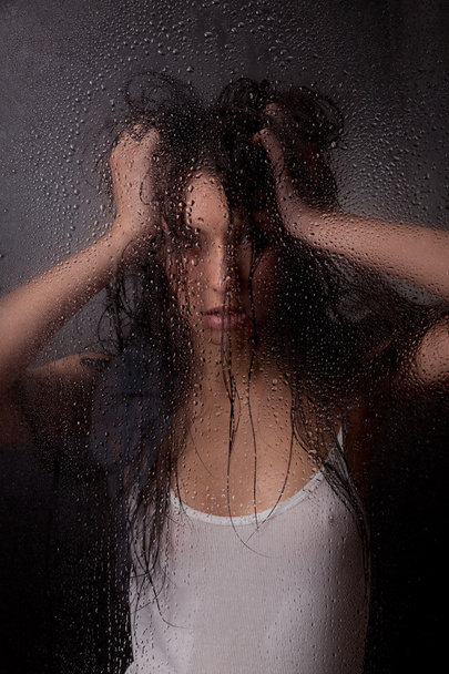 A beautiful girl in a white t-shirt, on a black background behind the glass. - Photo, image