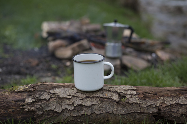 Mug stands on a log near the fire at a campsite. - Zdjęcie, obraz