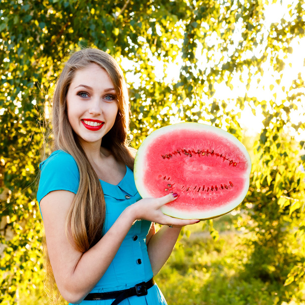 Beautiful blonde girl in a blue dress holds a half of watermelon - Fotoğraf, Görsel