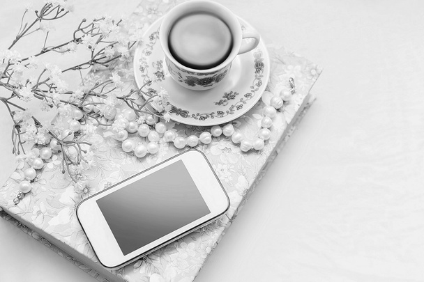 Phone with a cup of tea and pearl necklace with flowers on a white satin background. A book, tea cup, telephone and decoration. - Photo, Image