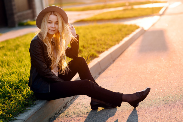 Young and beautiful girl in a hat sitting on the curb side of the road at sunset - Photo, Image