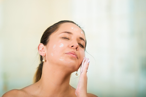 Pretty young healthy hispanic woman headshot with naked shoulders, applying cream to face using hands - Photo, image