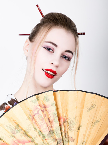 beautiful girl dressed as a geisha, she holds a chinese fan. Geisha makeup and hair dressed in a kimono. The concept of traditional Japanese values - Foto, Imagem