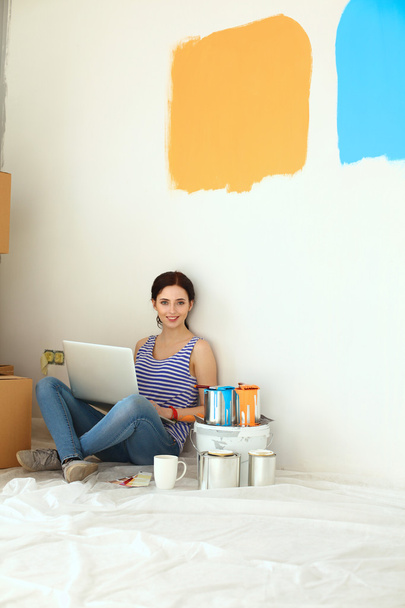 Πορτρέτο της νεαρής γυναίκας ενώ ζωγραφική νέο διαμέρισμα, κάθεται με τον φορητό υπολογιστή - Φωτογραφία, εικόνα