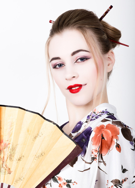 beautiful girl dressed as a geisha, she holds a chinese fan. Geisha makeup and hair dressed in a kimono. The concept of traditional Japanese values - Zdjęcie, obraz
