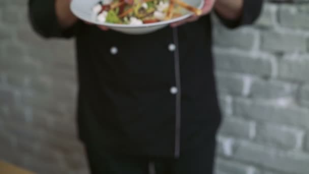 Closeup chef-kok in zwart uniform presenteert heerlijke salade op muur achtergrond 4k - Video