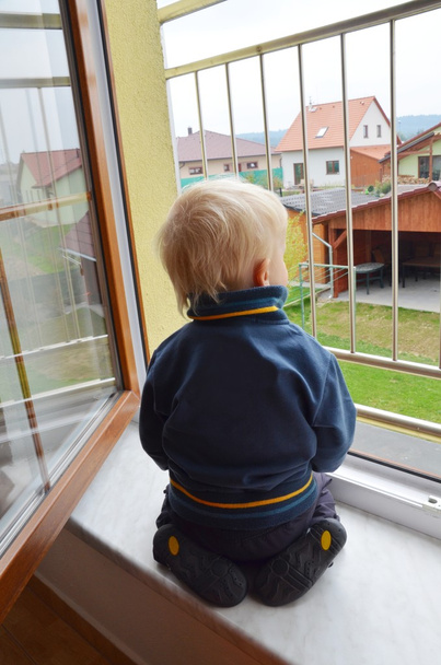 Słodki samotny chłopiec oglądający ogród przez okno. Przed nim jest barierka.. - Zdjęcie, obraz