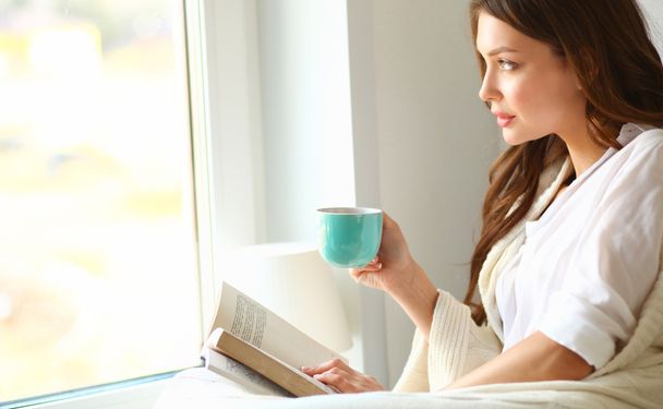 Young woman at home sitting near window relaxing in her living room reading book and drinking coffee or tea - Foto, imagen