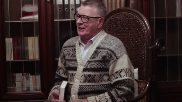 Old man with glasses cheerful grandfather remembers the youth on a rocking chair near the bookcase - Footage, Video