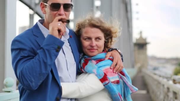 Woman hugs man smoking cigar and strokes his cheek on bridge - Filmati, video
