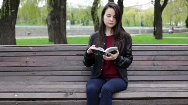 woman is sitting on the bench in the park and reading the book - Materiał filmowy, wideo