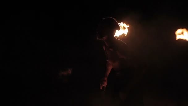 Fire show performance. Handsome male fire performer twirling fire baton and making fire breathing spitting flame against black background. Slow motion - Materiał filmowy, wideo