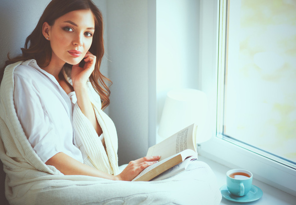 Молодая женщина дома сидит у окна, расслабляясь в своей гостиной, читает книги и пьет кофе или чай - Фото, изображение