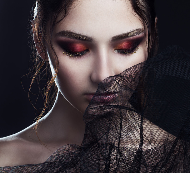 Portrait of mysterious beautiful brunette through black veil and posing over black background. Gold eyebrows, wet lips, red shadows. Close up. Vogue style. Studio shot. Haute couture concept.  - Foto, afbeelding