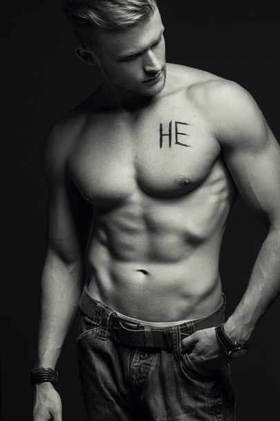 Handsome muscular male model with intense glance posing over grey background. Perfect body with the inscription "he" on his chest . Studio shot - Photo, Image