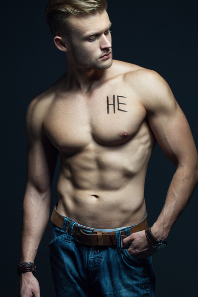 Handsome muscular male model with intense glance posing over grey background. Perfect body with the inscription "he" on his chest . Studio shot - Photo, Image