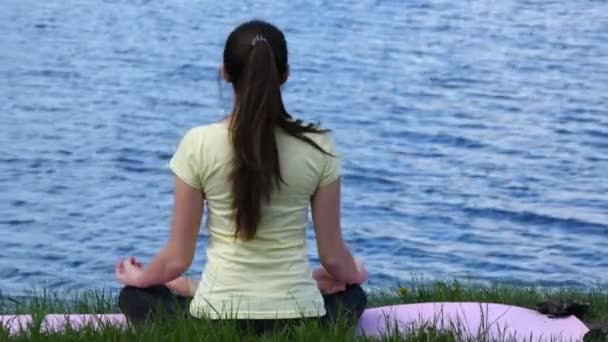 Fiatal nő, pihentető a strandon meditál. Lány ül a fűben és meditálni csinál jóga gyakorlatok során a lótusz helyzet folyó mellett - Felvétel, videó