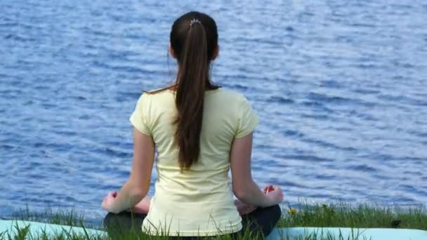 Young woman relaxing meditating on the beach. Girl sitting on the grass and meditate beside the river doing yoga exercises during lotus position - Footage, Video