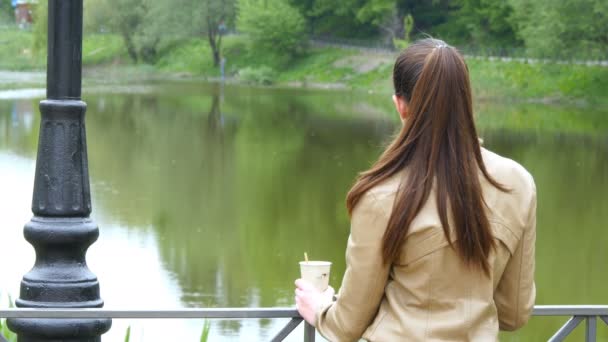 Vista posterior de la joven bebiendo taza de café para llevar de la taza desechable. Mujer cuidadosamente mira hacia el río y la celebración de la taza de té para calentar
 - Imágenes, Vídeo