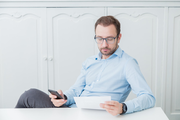 Бізнесмен сидить за своїм столом, дивлячись на паперовий документ і тримаючи смартфон
       - Фото, зображення