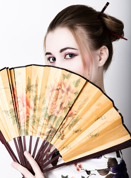 beautiful girl dressed as a geisha, she holds a chinese fan. Geisha makeup and hair dressed in a kimono. The concept of traditional Japanese values - Φωτογραφία, εικόνα