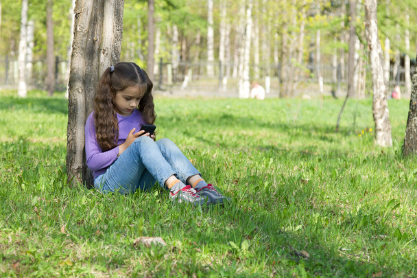 Cute little long-haired girl sitting on the grass in a park with a mobile phone in her hands and sending message on phone mobile - Zdjęcie, obraz