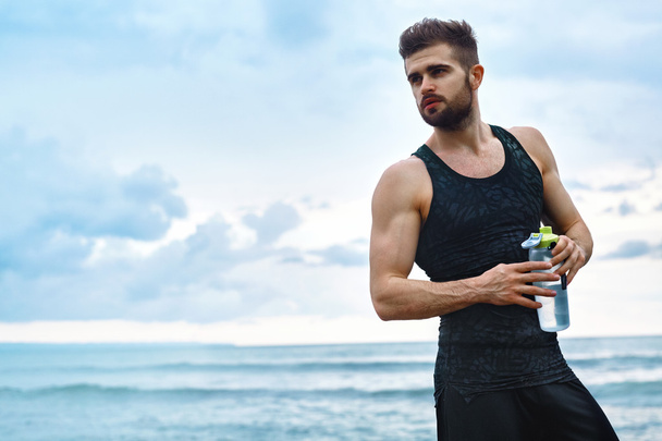 Фитнес-человек с водяной бутылкой отдыхает после тренировки на пляже
 - Фото, изображение