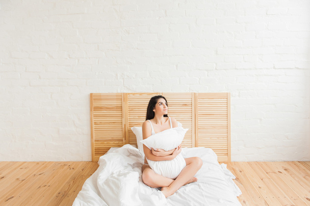 Smiling woman with pillow sitting in bed and looking up - Fotografie, Obrázek